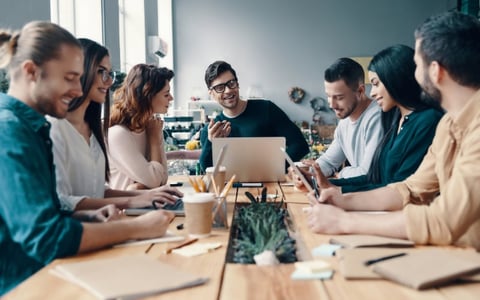 Diverse team of professionals gathered around a table with laptops and coffee, collaborating in a bright modern office setting