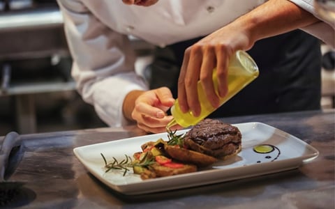 Chef in white jacket drizzling golden sauce over a plated steak with vegetables and herbs